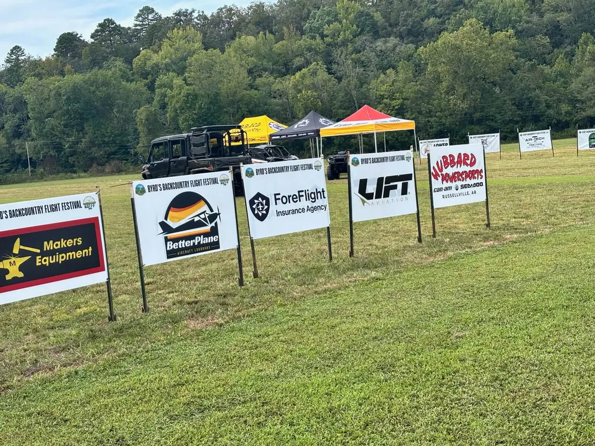 A row of sponsor signs on a grass field at Byrd's Backcountry Flight Festival, including the BetterPlane Aircraft Logbooks sign, with tents and utility vehicles and a wooded hill behind them.